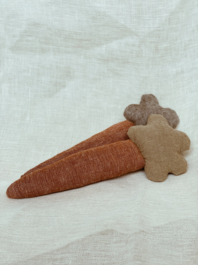 A head-on photo of two carrots rattles resting against a white linen background. The front carrot rattle has a sage-green carrot top. The back carrot rattle has a slightly darker, more olive-green carrot top. The carrot tops are circular, almost like little clouds.