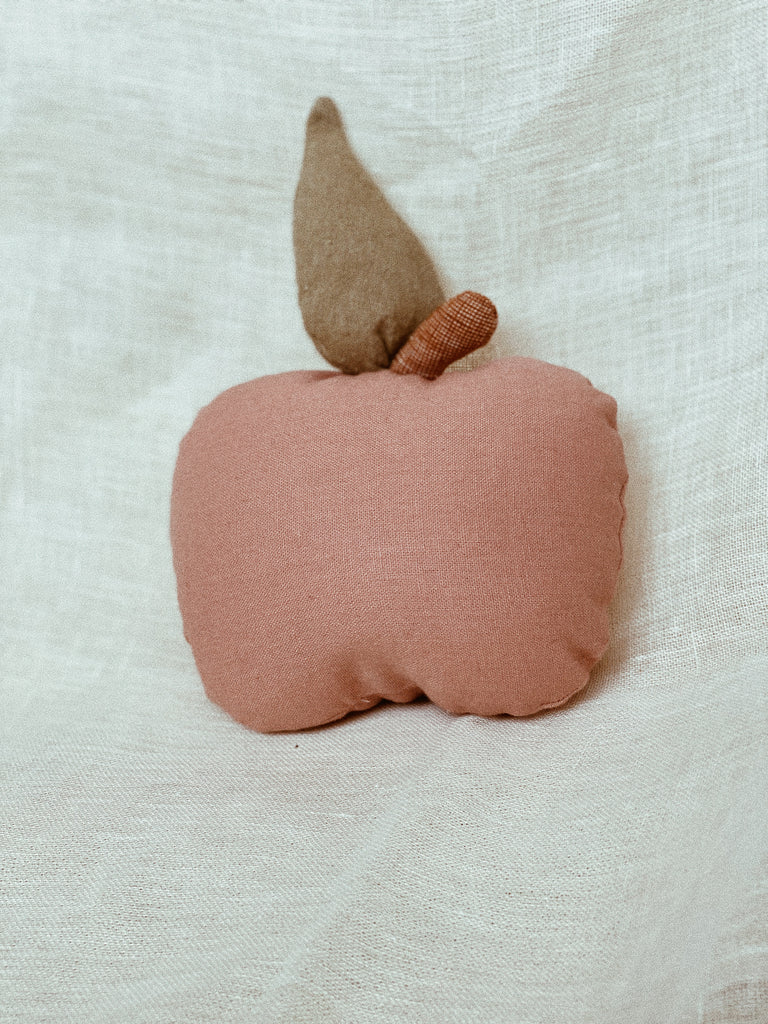 Apple rattle made out of dusty pink linen rests against a white linen background. The little green leaf is sticking up, and the little brown stem arcs to the right. The photo is taken from a low angle.